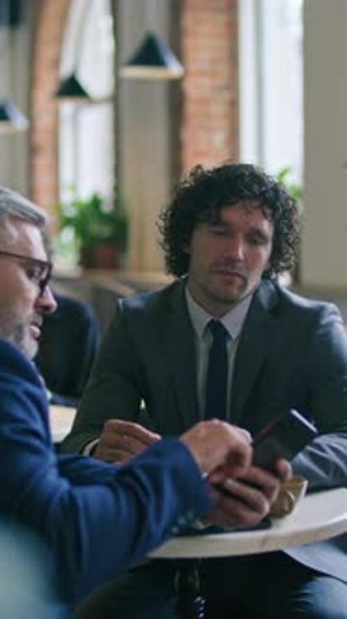 Businessman Explaining Information on Smartphone to Colleague at Cafe Meeting