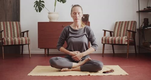 Jeune femme adulte caucasienne assise en position de lotus dans la pièce et méditant sur le mobilier de maison