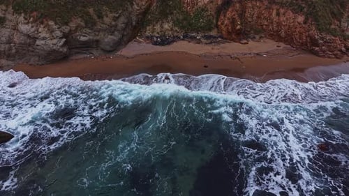 Waves crash on sandy shore beneath rocky cliffs at seaside location