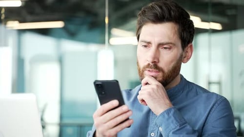 Thoughtful bearded businessman is using phone in business office. Serious manager works on a mobile