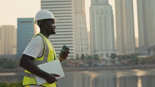 Portrait of Positive African American Engineer with Laptop Walking Along Embankment