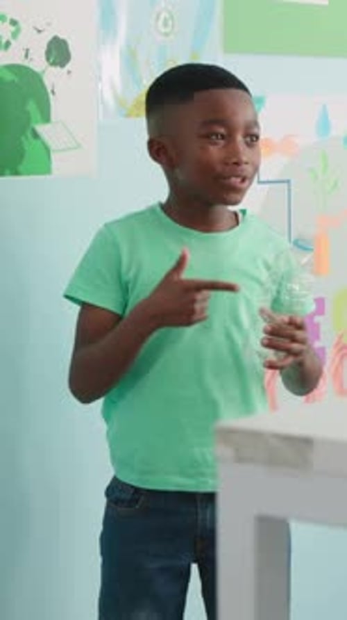 Vertical video: Reaching up at school, kid in green tee showing recycling, dropping bottle in bins