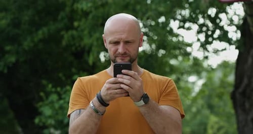 Young Attractive Bald Man with Bracelets and Tattoos on His Arm Uses the Phone to View News on