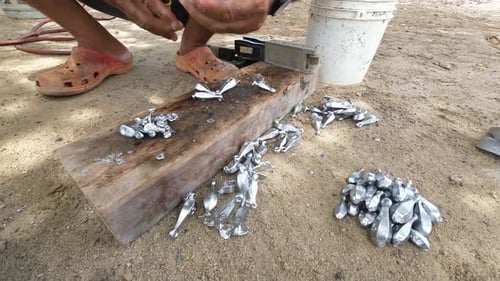 Person using pliers to handle hot molten metal poured into molds, on a wooden plank, outdoors, durin