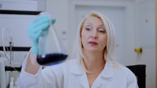 Woman in lab coat examines blue liquid in flask