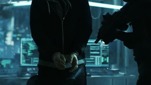 Un hacker masqué menotté est gardé par un soldat des forces spéciales entièrement armé. Ils sont à Hacke
