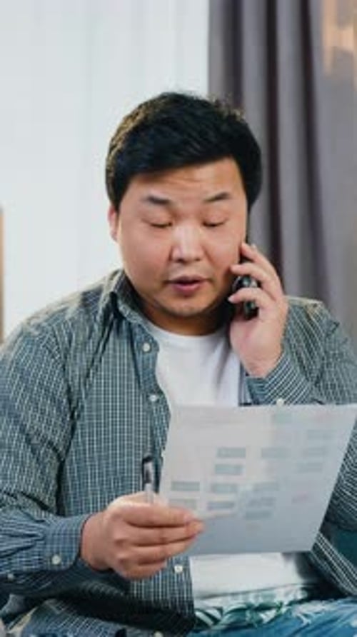 Man Sitting on the Couch at Home and Talking on Phone Discussing Data's From Report with Coworker