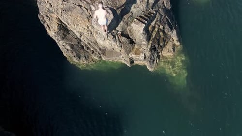 Drohnenaufnahme eines Mannes, der einen Rückwärtssalto auf einer Klippe ins Meer macht.