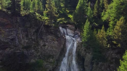 alpine waterfall on a rocky wall with pine forest, aerial shot at sunset