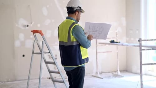 Engineer in Helmet Pointing Index Finger to Shabby Wall Holding Drawing Paper in Hand