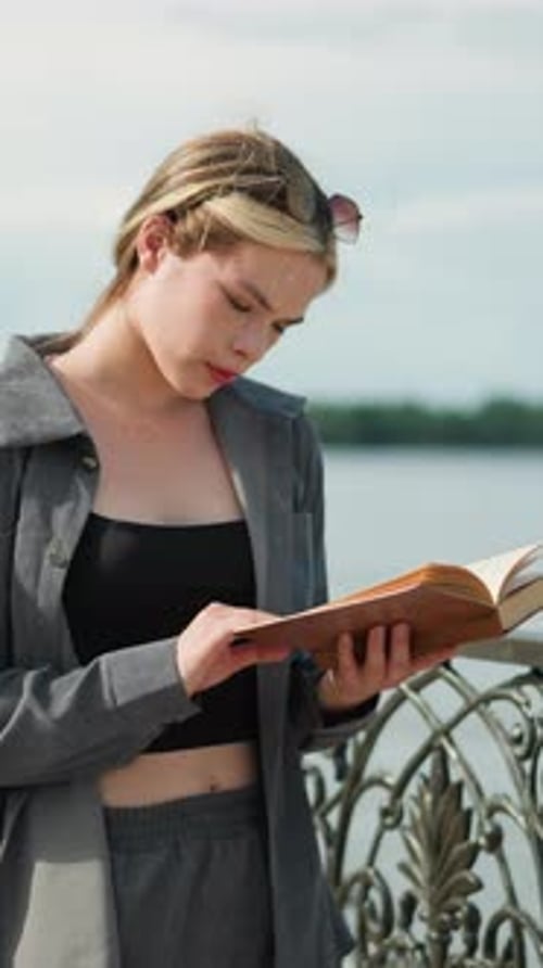 Focused Young Lady in Grey Reading Book As Wind Blows Pages By River