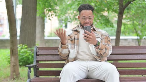 Young Multi Ethnic Man Doing Video Chat on a Phone, Sitting Outdoor on Bench