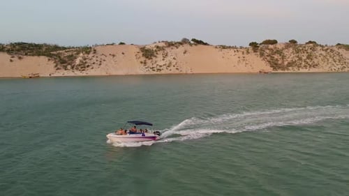 People Ride On Speedboat Leaving Wake In The River With Sand Dunes In The Background. - aerial
