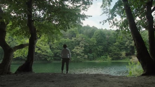 A Man standing in front of the lake in a forest with the trees and blue sky