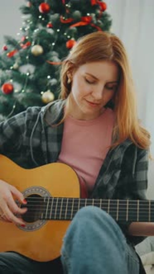Woman plays guitar by decorated Christmas tree indoors