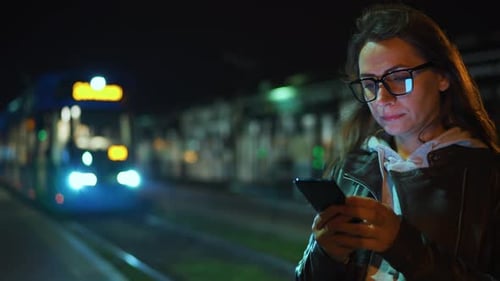 Frau steht nachts mit Smartphone an einer Haltestelle und wartet auf die Straßenbahn