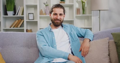 Joli jeune homme souriant et heureux assis sur un canapé confortable à la maison et regardant la caméra