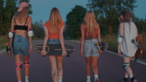 Stylish Girls Walk Along Country Road Holding Skateboards