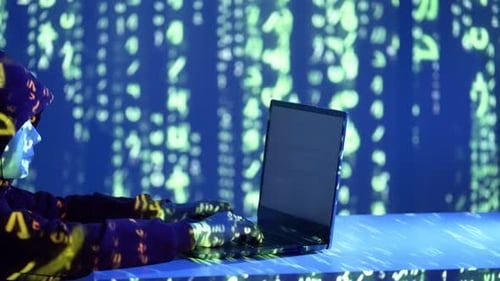 Hacker en sweat à capuche avec masque en train de taper sur un ordinateur portable dans une pièce sombre avec projection de code vert
