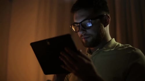 Young man in glasses uses tablet pc for social networking late at night