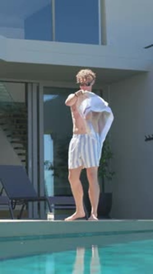 Vertical video: Exiting sliding glass door, man in striped trunks laying towel at poolside to rest