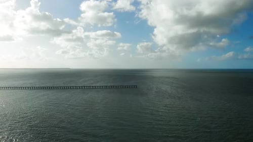 A long, wooden pier sits in the deep, dark, blue water in the distance whilst we sweep from its edge