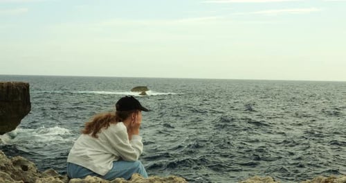 Teenage Girl Contemplates Rolling Waves Sitting Alone on Rock at Sea Resort