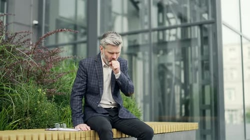 Sick bearded business man employee coughing a lot while sitting on bench near modern business centre