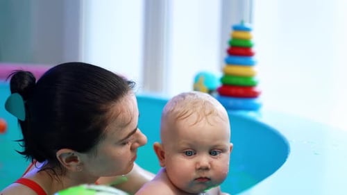 Woman is teaching a little baby boy to swim. Happy child splashes his hands by the water and smiles.