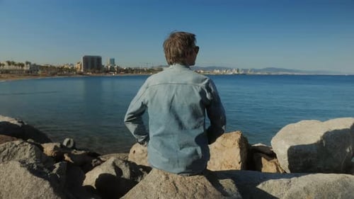 A Man is Peacefully Seated By the Ocean on Rocks Enjoying the Serene Seaside View