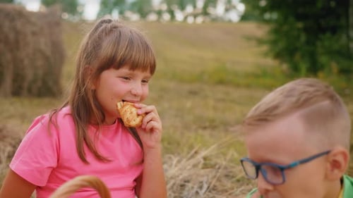 Little Girl Enjoying Snacks Outdoors with Boy Seated Nearby in Open Field