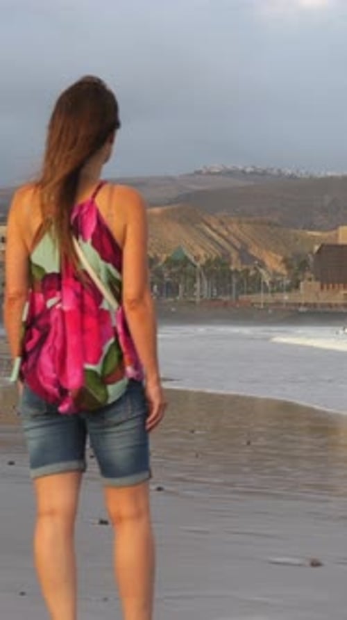 Mature woman standing on the shore, gazing at the sea and the coastal city in the distance