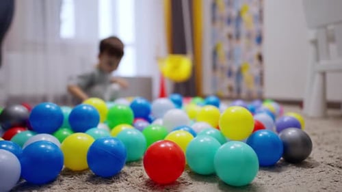 Multiple colorful balls are on the floor of the room. Little kid falls on the balls smiling happily.