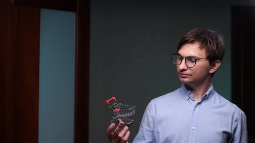 Empleado de oficina con un pequeño carrito de compras reflexionando sobre las compras y el consumismo