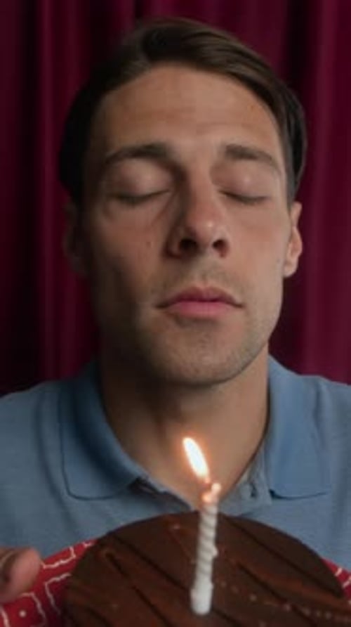 Vertical of Young Man Making Wish and Blowing Candle on Birthday Cake