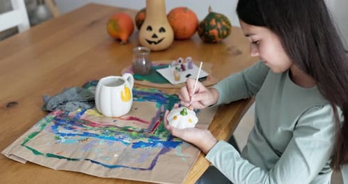 Female child painting pumpkin cup at home for Halloween party - Holiday, art and fall season concept