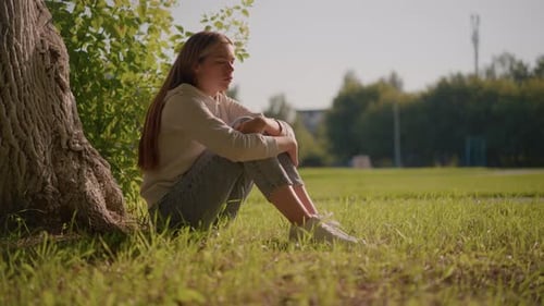 Thoughtful Woman Sitting on Grass in Sunlight with Urban Stadium Background
