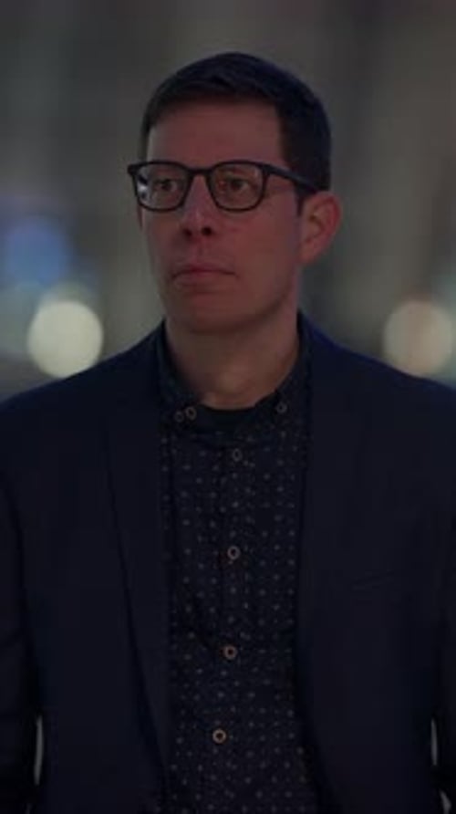 A Thoughtful Man Wearing Glasses Stands Against a Backdrop of Vibrant City Lights