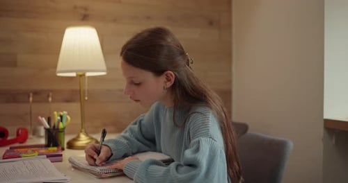 Teenage Girl in Sweater Doing Homework and Writing in Her Notebook Sitting at Table with Desk Lamp