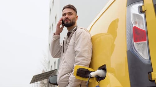 Young Man Speaks on the Phone While the Electric Car is Charging Outdoors