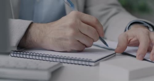 Closeup of Male Hands Writing in Notebook While Businessman Working at Desk in Office