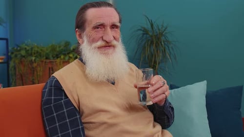 Durstiger älterer Mann sitzt zu Hause mit einem Glas natürlichem Aqua und trinkt stilles Wasser