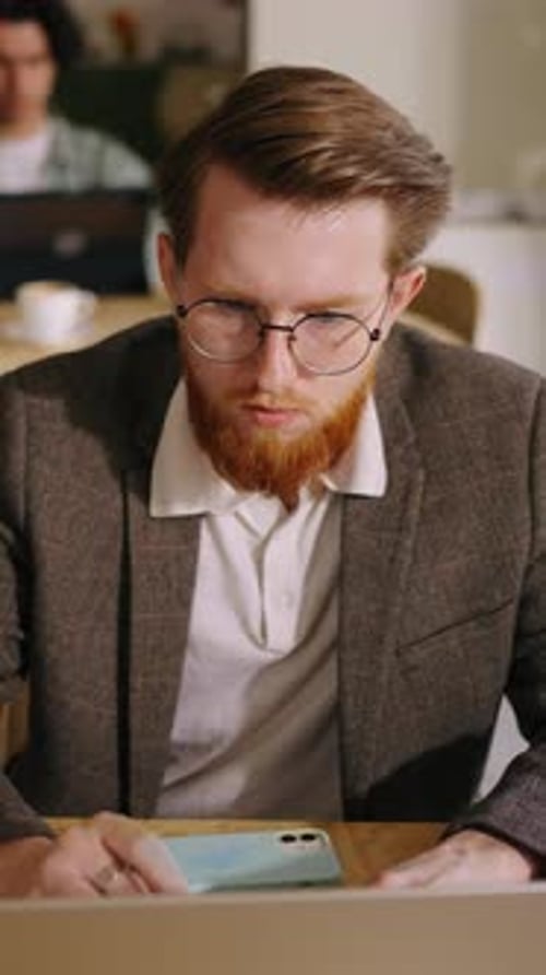 Jeune professionnel concentré avec barbe rousse et lunettes travaillant sur un ordinateur portable dans un espace de travail partagé confortable