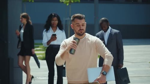 A Young Professional in a Suit Walks in a Modern Area with a Laptop and Coffee Showing Ambition