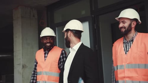 Ein fröhliches Team von Ingenieuren in weißen Schutzhelmen verlässt die unvollendete Konstruktion und lächelt zu