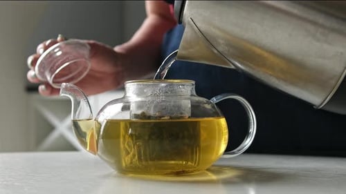 Girl Pouring Hot Water From The Kettle Into Teapot With Green Tea In The Kitchen