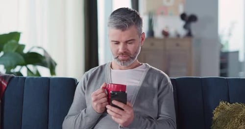 Man Sits on Couch, Drinks From Mug, Uses Phone