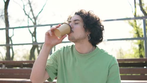 Close Up Shot of Young Curly Brunette Man Sitting in Park Drinking Morning Coffee