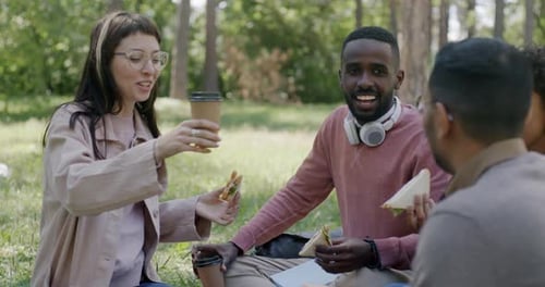 Multiethnic Group of Students Clinking Coffee Cups and Eating Sandwiches Having Picnic in Park