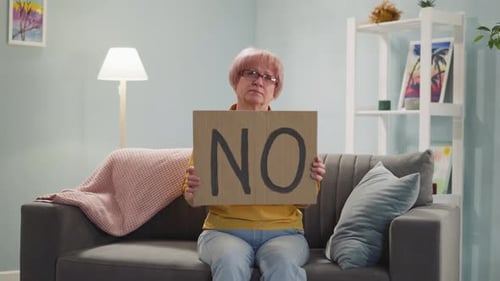 Une femme âgée bouleversée tient une pancarte avec le mot « NON » sur le canapé
